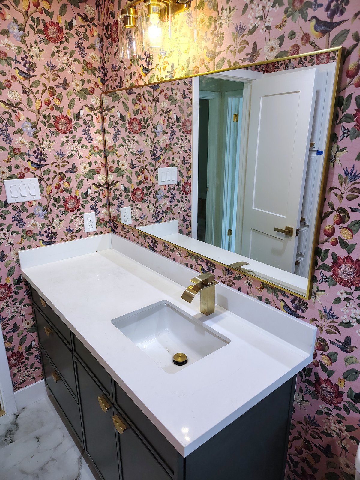 Dark green vanity with a white quartz top, a gold-framed mirror and pink floral bird wallpaper.