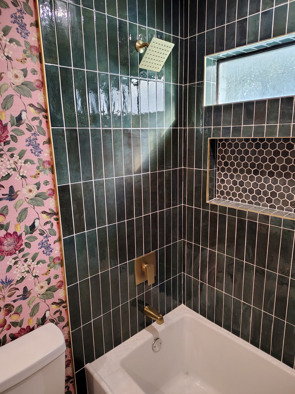 Close-up of the green vertical tile shower with a gold rain shower head and a hex-tile niche beside floral wallpaper.