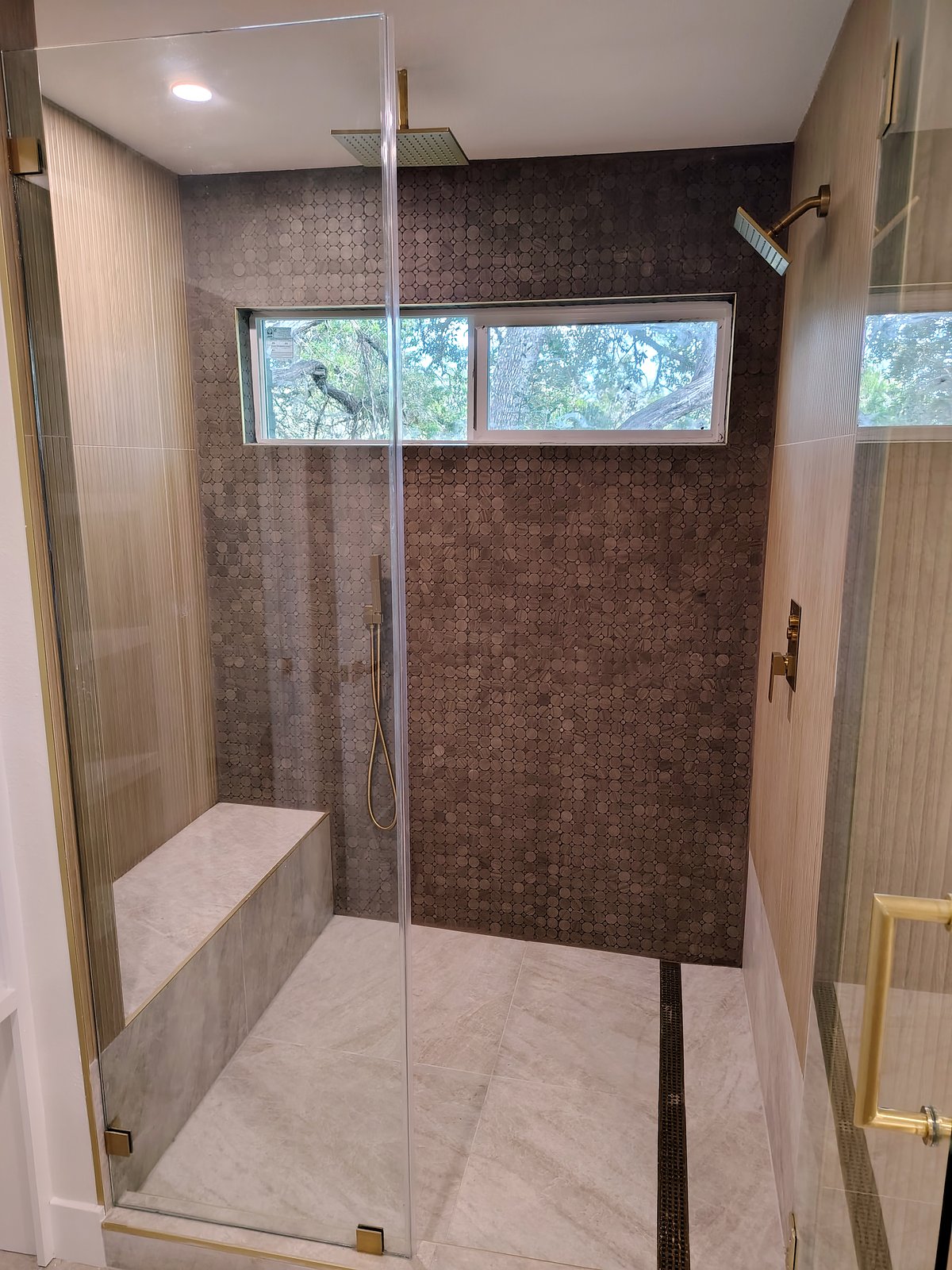 Curbless walk-in shower with a built-in bench, brown penny-round tile wall, wood-look tile, linear drain and frameless glass.