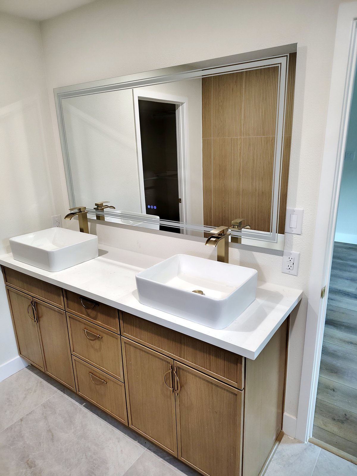 Wood double vanity with white vessel sinks, brushed-gold faucets and pulls, and a large backlit mirror.