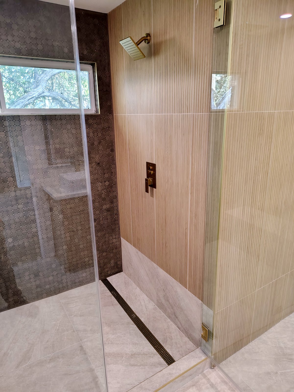 Shower with a brushed-gold valve and shower head, wood-look wall tile, penny-round tile and a linear floor drain.